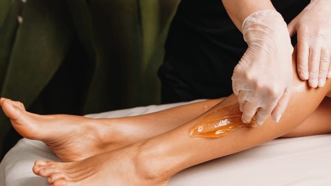 Gloved hands spreading sugar paste on a woman's lower leg as she lies on a treatment couch